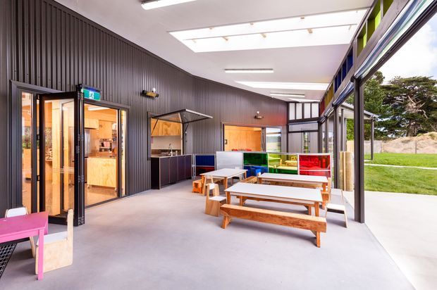 Bright contemporary communal interior with an open-plan dining and breakout area. The space features dark vertically ribbed wall cladding, polished concrete flooring, extensive black-framed glazing, skylights, colourful built-in cabinetry, and timber picnic-style tables and benches. A kitchen or service area is visible through the doorway, with landscaped grounds beyond the glazing.