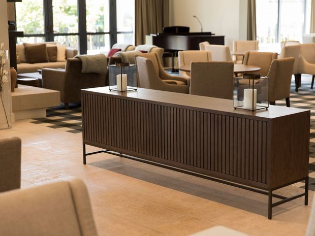 A refined hotel or hospitality lounge with a long dark timber slatted console cabinet in the foreground, surrounded by upholstered lounge chairs, sofas, side tables, candle lanterns, and a black grand piano. Large windows with curtains provide abundant natural light, while patterned flooring and warm neutral finishes create an elegant, welcoming atmosphere.