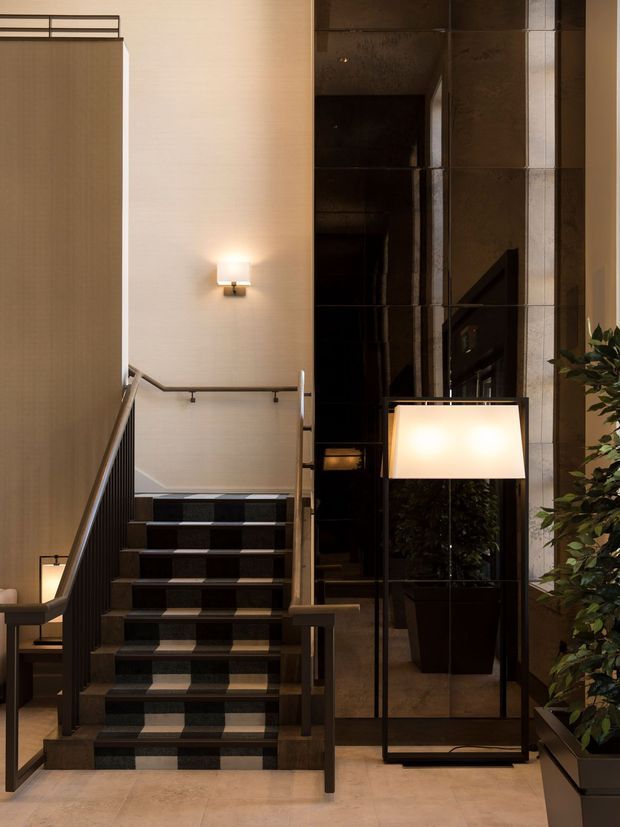 A refined interior stair hall with a straight staircase featuring dark patterned carpeted treads, dark timber or metal handrails, and a pale textured wall. A tall mirrored or dark reflective wall panel sits beside the stairs, accompanied by a large floor lamp and indoor planting. The space has warm layered lighting, neutral stone-toned flooring, and a polished contemporary hotel-like atmosphere.