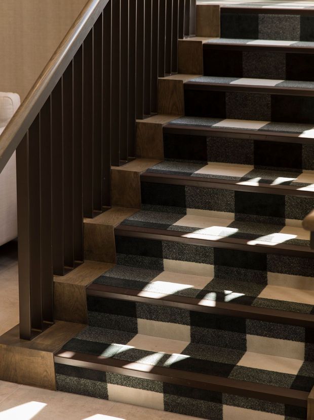 Close-up interior view of a contemporary stairway with dark brown timber or wood-look vertical balustrade and side detailing. The stair treads and risers feature a graphic checkerboard-like combination of dark textured surfaces and pale stone or tile, with warm timber side elements and strong sunlight casting shadows across the steps.