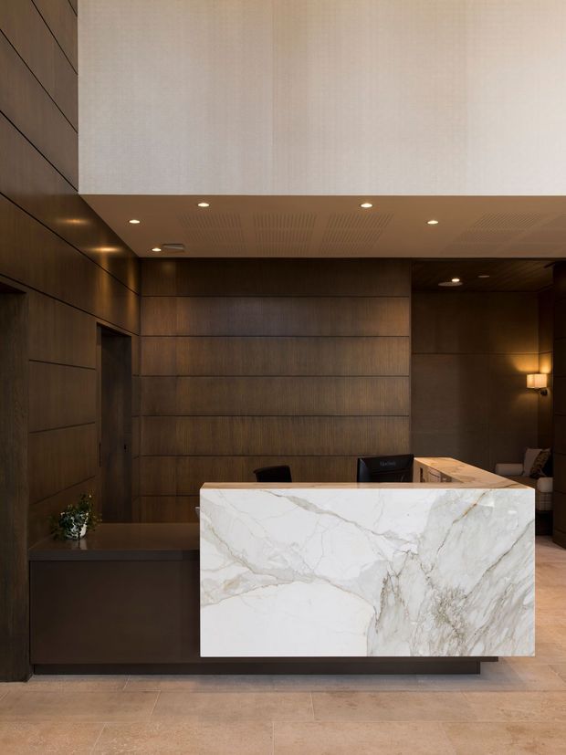A refined reception or front desk area with dark timber wall panelling, a large white veined stone reception counter, warm recessed lighting, and pale stone flooring. The space has a calm, luxurious commercial hospitality feel with a high ceiling and concealed lighting.