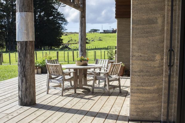 Sunlit covered outdoor deck with a timber dining table and chairs beside a textured earthen wall and dark-framed door. Rustic timber posts support the shelter, while a fenced green rural landscape is visible beyond.