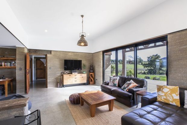 A bright contemporary living room with polished concrete flooring, textured light brick walls, expansive dark-framed glazing, and a large white ceiling. The room includes black leather sofas, a timber coffee table, woven rug, timber media cabinetry, open shelving, and an industrial-style pendant light. A rural landscape is visible through the windows.