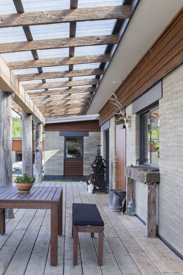 A covered outdoor terrace or courtyard with a timber plank deck, substantial rough-hewn timber posts and beams, and a translucent roof canopy. The space includes a wooden dining table and bench, a weathered timber-clad wall, grey textured masonry, dark-framed windows and door, rustic metal objects, and a potted succulent. The architecture blends rugged natural materials with contemporary detailing.