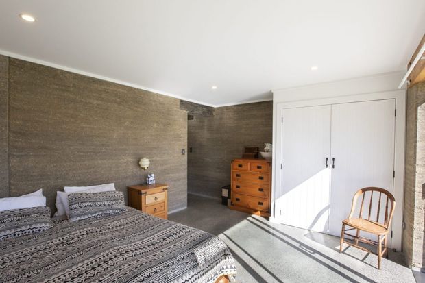 Bright bedroom with a large bed dressed in patterned grey bedding, textured brown-grey wall finishes, white built-in double wardrobe doors, timber chests of drawers, and a wooden chair. Recessed ceiling lights and strong sunlight create a warm, inviting atmosphere.