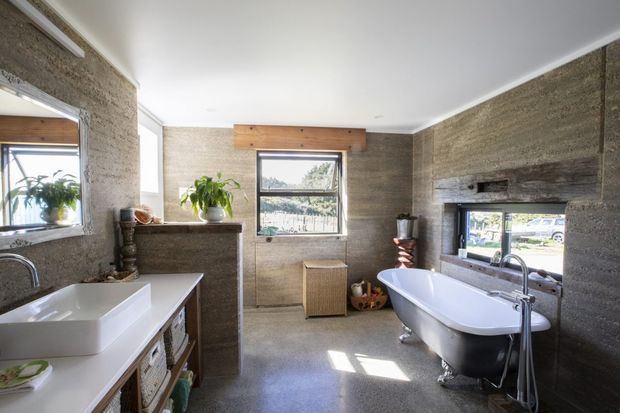 A bright, spacious bathroom with textured earthy grey-brown wall finishes, a freestanding white clawfoot bathtub, and a white vessel sink on a timber vanity. Multiple windows provide natural light and views outside, while open shelving and potted plants add warmth and an organic character.
