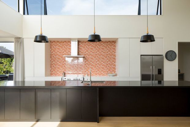 A contemporary kitchen with a large dark island, white full-height cabinetry, stainless-steel appliances, and a distinctive orange patterned splashback. Three black pendant lights hang above the island, while high-level glazing and a double-height ceiling bring in abundant natural light.
