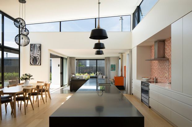 A bright, open-plan contemporary kitchen and dining area with a large central island, integrated cabinetry, stainless-steel appliances, and a textured warm-toned splashback. The space has pale timber flooring, white walls and joinery, extensive black-framed glazing, and a high ceiling with clerestory windows. A dining table sits to the left, while the kitchen opens toward a living area at the rear. Multiple pendant lights provide visual focus over the island and circulation zone.