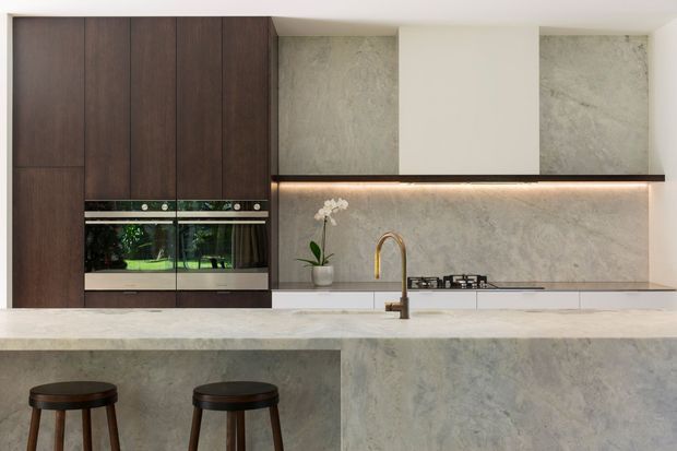 Contemporary minimalist kitchen with a long island, integrated dark timber cabinetry, built-in double ovens, pale stone-look benchtops and splashback, brass-toned tapware, gas cooktop, and warm under-cabinet lighting. Two stools sit at the island, with a small orchid adding understated decor.