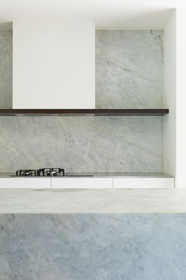 Minimalist kitchen composition featuring a large pale grey veined stone backsplash and matching waterfall island. White handleless cabinetry and a simple gas cooktop sit beneath a dark timber shelf, creating a restrained, monochromatic interior with subtle natural texture.