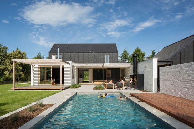 Exterior view of a modern multi-volume home arranged around a long swimming pool. The house has dark standing-seam pitched roofs, grey and white horizontal cladding, large glazed openings, covered outdoor areas, a timber pool deck, and landscaped lawn. The composition highlights the pool and rear facade under a bright blue sky.