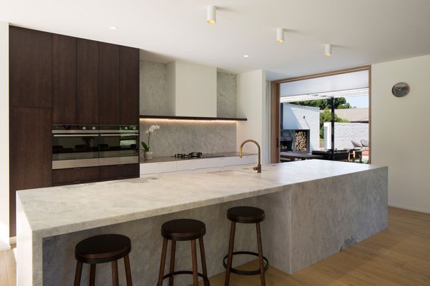 Contemporary open-plan kitchen dominated by a large stone island with seating for three. Dark timber full-height cabinetry incorporates built-in ovens, while a pale textured stone backsplash and matching island create a refined neutral material palette. The room opens toward an outdoor area through large glazed doors, with warm timber flooring and understated ceiling lighting.