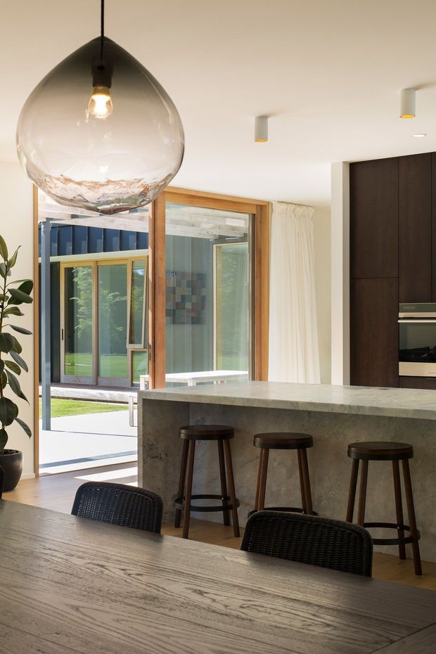 A bright contemporary kitchen and dining area with a large grey stone island and three dark timber bar stools. A timber-framed glazed opening connects to an outdoor courtyard, while dark full-height cabinetry houses integrated appliances. The room features a large clear glass pendant light, recessed ceiling spotlights, pale timber flooring, a timber dining table, woven dining chairs, and white curtains.