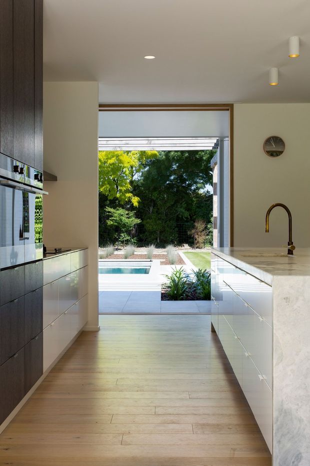 A contemporary galley kitchen viewed toward a large open glazed doorway leading to a landscaped courtyard and swimming pool. The kitchen has dark wood-toned tall cabinetry on the left, pale glossy lower cabinetry, a light stone island and benchtop with an integrated sink and curved brass-toned tap, recessed and cylindrical ceiling lights, and light timber flooring. The space is bright and visually connected to the outdoor area.
