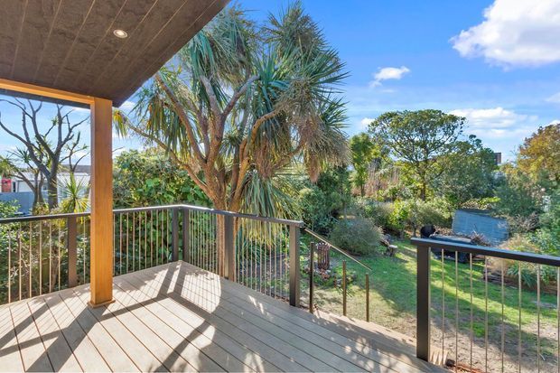 A sunny elevated timber deck overlooking a lush landscaped garden, with dark metal balustrades, a timber post and covered roof soffit. The scene includes established tropical-style planting, bright blue sky and neighbouring residential structures in the background.