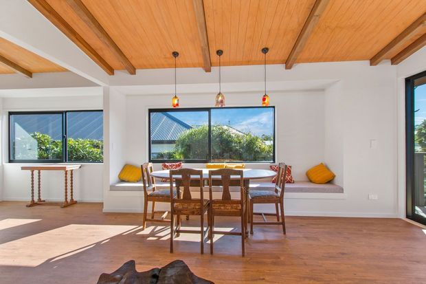 Bright dining area with a timber-lined ceiling, exposed timber beams, white walls, warm timber flooring, and a built-in window seat. A wooden dining table and chairs sit in front of large windows, with colourful pendant lights and cushions adding small accents. The space has a relaxed, warm, contemporary character.