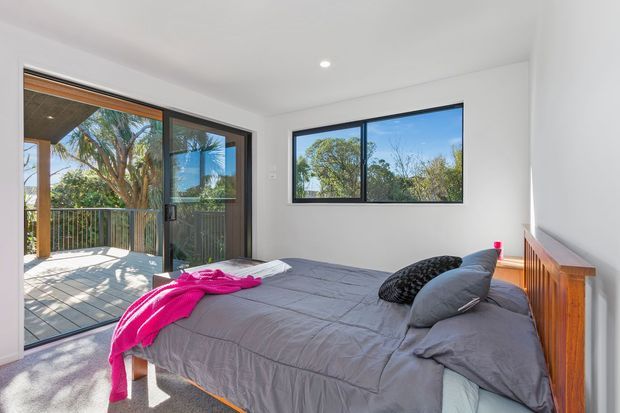 Bright contemporary bedroom with a grey upholstered bed, layered grey bedding, and a vivid pink throw. White walls and ceiling, dark-framed glazing, and a timber headboard create a clean, modern interior. Sliding glass doors open to a timber balcony overlooking leafy vegetation, with additional horizontal window glazing bringing in abundant daylight.