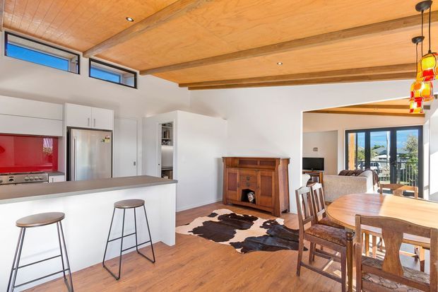 Open-plan residential kitchen, dining, and living area with white cabinetry, stainless-steel appliances, a dark benchtop, timber flooring, and a prominent timber-lined ceiling with exposed beams. A timber dining table and chairs occupy the foreground, while a traditional timber cabinet and cowhide rug add warmth. Large glazed doors connect to an outdoor balcony or terrace.