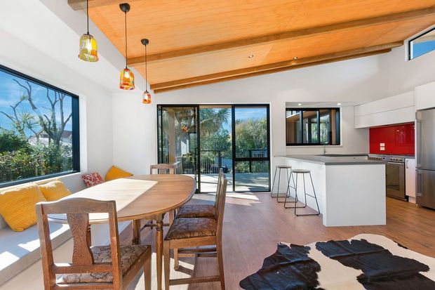 Open-plan kitchen and dining area with a warm timber-lined ceiling, exposed timber beams, white walls, timber flooring, and extensive glazing. A round timber dining table sits beside a built-in window seat, while the contemporary kitchen features a white island, stainless-steel appliances, and a vivid red splashback. Sliding glass doors open to an outdoor setting.