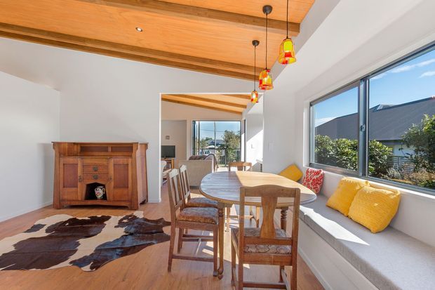 Bright, warm dining area with a timber-lined ceiling featuring exposed beams, white walls, timber flooring, and a built-in window seat beneath large glazing. A round timber dining table with wooden chairs sits beside the window, with colourful pendant lights overhead. A vintage timber cabinet and cowhide rug add rustic character, while adjacent living space is visible beyond.