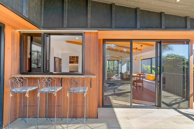Exterior view of a contemporary home entertaining area with a built-in timber-clad outdoor bar, metal bar stools, and large sliding glazed doors opening into a bright living and dining space. The facade combines warm timber framing with dark vertical metal cladding and extensive glazing, with a paved terrace in front.