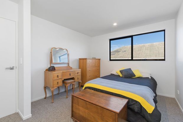 A compact bedroom with white walls and ceiling, grey carpet, and a single bed dressed in layered grey, blue, and yellow bedding. A timber vintage-style dressing table with an arched mirror and matching tall chest of drawers sits beside the bed, while a timber blanket chest is positioned at the foot. A black-framed horizontal window provides natural light, with a neighbouring tiled roof visible outside.