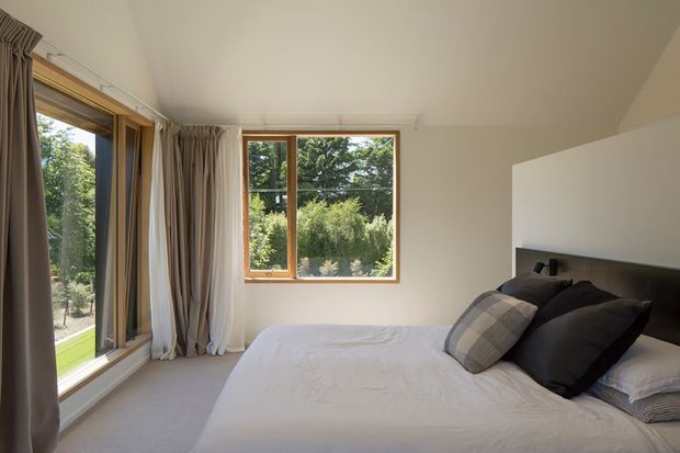 A bright, calm bedroom viewed from inside, with a large bed, dark upholstered headboard, layered neutral bedding and cushions, pale carpet, and warm timber-framed windows. Full-height glazing and a second window overlook leafy gardens, while beige curtains frame the openings. The room has a simple contemporary fit-out with clean lines and a light neutral palette.