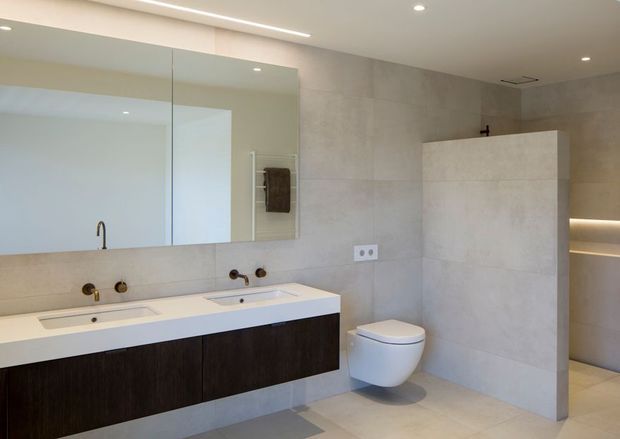 A spacious, contemporary bathroom with a double vanity, wall-mounted toilet, large frameless mirror, and walk-in shower enclosure. The room features pale stone-look wall and floor tiles, a dark timber vanity, recessed ceiling lighting, and softly illuminated recessed shelving.