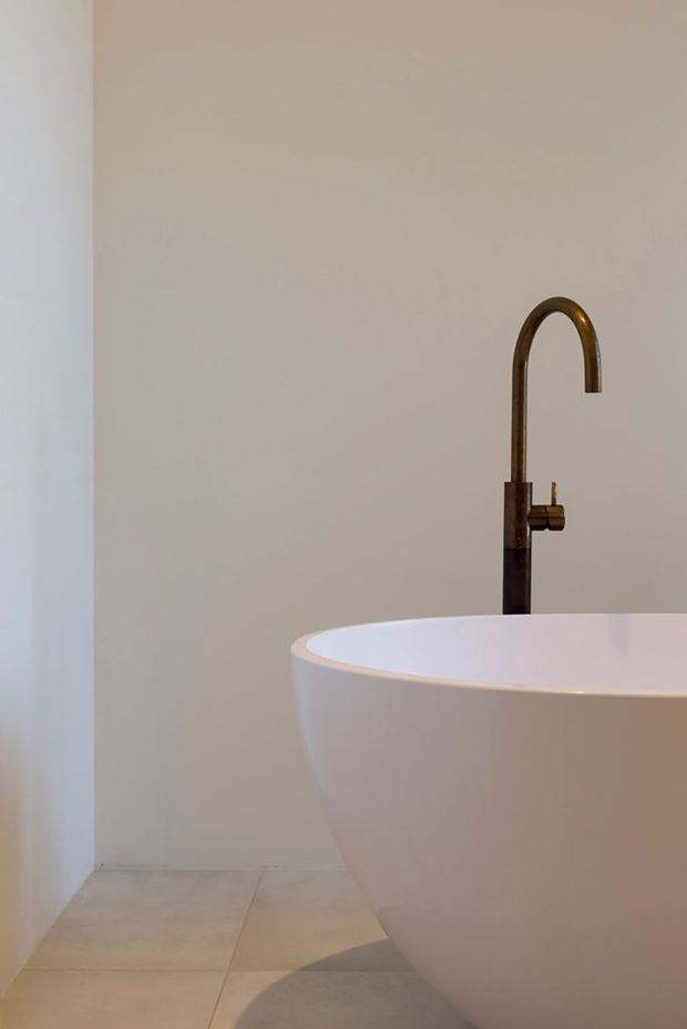 A minimalist bathroom detail featuring a freestanding white oval bathtub and a tall dark bronze or black gooseneck wall-mounted tap. The room has warm off-white plaster walls, pale neutral floor tiles, and a clean, uncluttered composition with soft natural light.