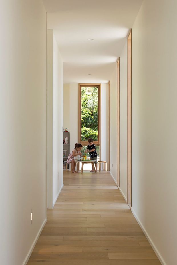 A long, bright residential hallway with pale painted walls, light timber flooring, and full-height timber-framed built-in cabinetry or doors along the right wall. The corridor leads to a sunlit sitting area with a narrow window framing greenery, where two children are seated at a small table. The design is clean and understated, with recessed ceiling lights and minimal detailing.