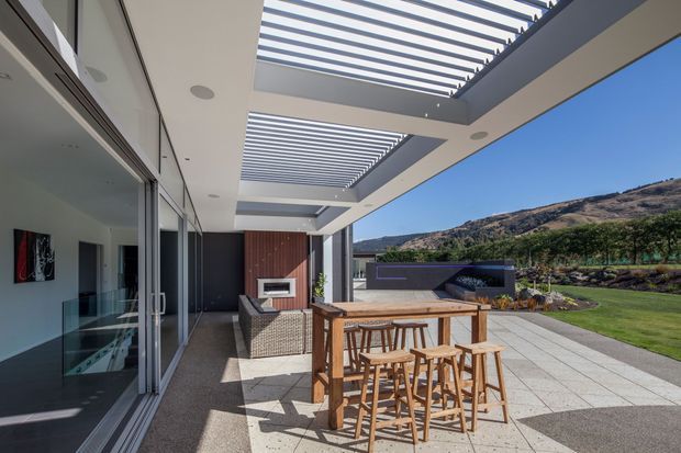 A contemporary outdoor entertaining terrace beside a glazed living area, featuring a timber bar-height dining table with stools, wicker seating, a built-in outdoor fireplace or barbecue set into a dark feature wall, and a large paved terrace beneath a slatted pergola. The scene opens onto a manicured lawn, landscaped planting, and a dry mountain or hillside setting under clear blue skies.