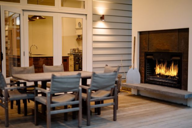 A warm, covered outdoor living and dining area with a timber dining table and chairs, beside a large built-in fireplace with a live fire. White horizontal weatherboard cladding, pale timber decking, and glazed doors connect the space to an interior kitchen. The setting feels cozy and inviting, with a relaxed coastal or traditional character.