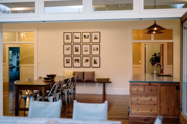 A warm, open-plan dining and kitchen area with a dark timber dining table and mixed chairs in the foreground, a built-in bench seat beneath a grid of framed artwork, and a timber kitchen island with a dark stone benchtop to the right. High-level clerestory windows and internal glazed openings bring in natural light, while a black pendant light hangs above the island. The space has timber flooring, light painted walls, and a relaxed contemporary-traditional character.