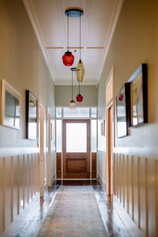 A long, narrow residential hallway leading to a timber front door with frosted glass sidelights and transom glazing. The corridor has white painted wainscoting, pale green walls, framed artwork, polished stone or tile flooring, and a decorative white paneled ceiling with multiple colourful pendant lights. Bright natural light enters from the glazed entry.