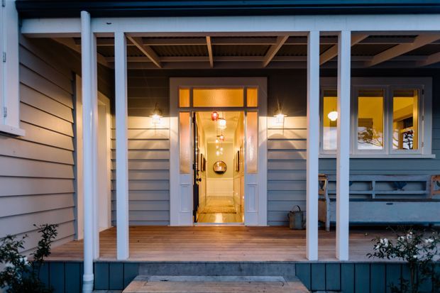 Front entry of a single-storey home viewed from a covered timber porch. The facade features grey horizontal weatherboard cladding, white-painted posts and trims, a glazed front door with warm interior lighting, and adjacent windows. A timber deck and concrete step lead to the entrance.