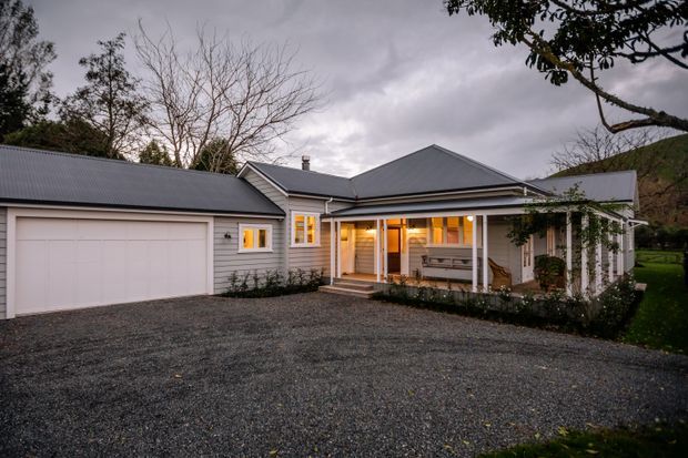 Exterior view of a single-storey grey-clad home at dusk, featuring a broad gabled roof, attached double garage, covered front porch, warm-lit windows, and a gravel driveway surrounded by mature landscaping.