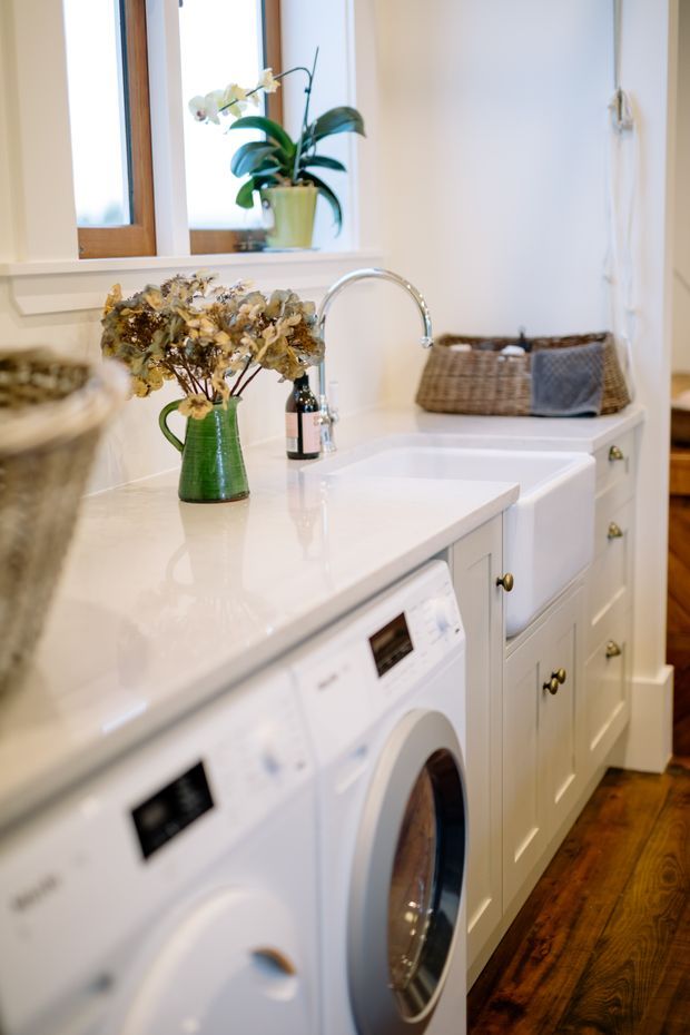 Bright, traditional-style laundry room with white built-in cabinetry, a glossy pale benchtop, integrated front-loading washing appliances, a deep utility sink with gooseneck tap, timber-framed windows, woven baskets, and warm timber flooring. Decorative flowers and an orchid add a homely touch.