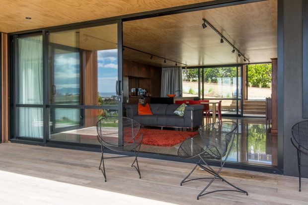 A modern indoor-outdoor living area viewed from a covered deck or terrace. Full-height black-framed sliding glass doors open onto a living room with a grey sofa, red rug and cushions, timber cabinetry, and a small dining area. The space features a warm timber-lined ceiling, dark structural framing, track lighting, polished reflective flooring inside, and outdoor wire lounge chairs in the foreground. Greenery and a residential landscape are visible beyond the glazing.