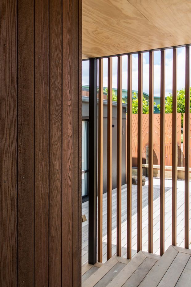 A close architectural view of a contemporary timber deck or covered outdoor transition space. Dark vertical timber wall cladding occupies the left side, while slim vertical dark metal or timber screening frames an open deck beyond. The ceiling and decking are timber, with a neighbouring structure and planted courtyard visible through the screen. The image emphasizes warm natural materials, strong vertical rhythm, filtered daylight, and privacy screening.