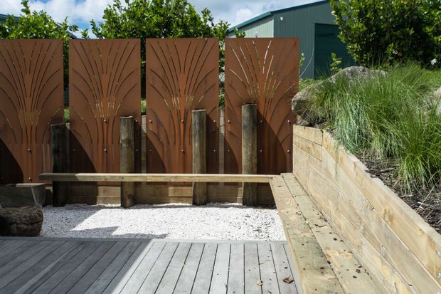 A landscaped outdoor courtyard or deck area featuring a grey timber deck, white gravel, a built-in timber bench, corten-toned decorative metal fence panels with organic cut-out patterns, and a timber retaining wall planted with ornamental grasses. A corrugated metal-clad building is partially visible behind the garden.