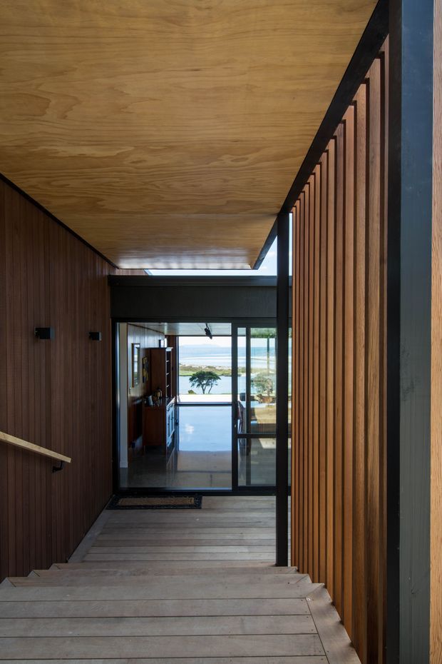 A narrow, modern timber-lined stairway and hallway descending toward a glazed entrance or living area. Warm wood panelling covers the ceiling and walls, with dark metal framing and vertical timber slats along the right side. Full-height glazing at the end opens views toward a bright coastal or waterfront landscape. The space has a restrained, warm architectural character with strong linear perspective.