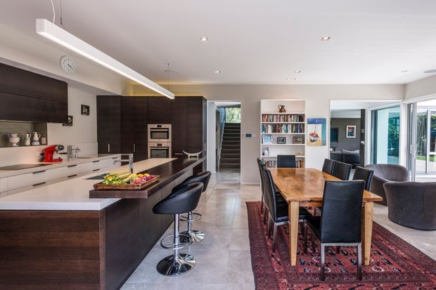 Open-plan contemporary kitchen and dining area with dark timber cabinetry, a large stone island with bar seating, integrated appliances, a timber dining table, built-in shelving, polished concrete flooring, and extensive glazing opening toward an outdoor area.