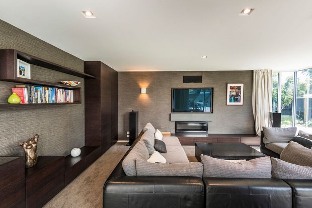 A contemporary living room viewed from behind a large sectional sofa, featuring dark built-in timber joinery and shelving along the left wall, textured grey wall finishes, a wall-mounted television with low cabinetry, recessed ceiling lights, and expansive glazing with curtains on the right. The room has a refined, neutral-toned interior with warm timber and leather accents.