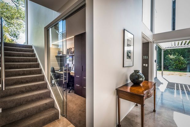 Contemporary residential entry and circulation space with a carpeted staircase, glass-fronted home office, console table, and strong visual connection to a bright covered outdoor area. The interior features clean white walls, dark timber joinery, tiled flooring, and extensive glazing.