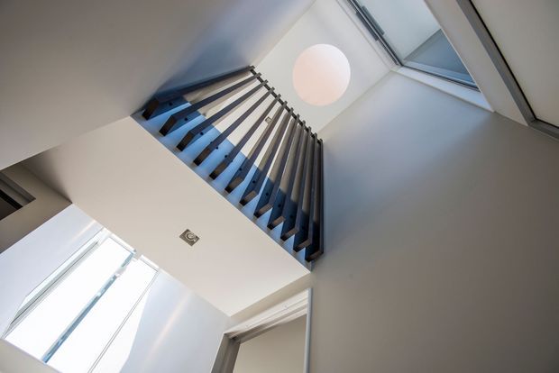 Upward view of a modern stairwell with dark vertical balustrade rails, white walls, roof glazing/skylights, and a spherical pendant light illuminating the double-height circulation space.