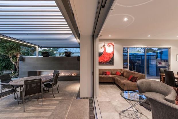 A contemporary indoor-outdoor living area viewed through fully opening glazed doors. The covered terrace has a dining table, outdoor seating, a long built-in fireplace, and a louvred pergola roof. Inside is a spacious living room with a sectional sofa, armchair, glass coffee table, dining area, tiled flooring, recessed lighting, and large windows.
