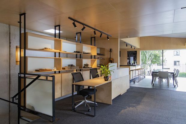 A contemporary open-plan commercial interior with a reception or hospitality counter, integrated shelving and display joinery, workstations, and a small dining or meeting area by large windows. The space features warm timber ceilings and cabinetry, black metal framing and track lighting, white counters, and dark carpet flooring.