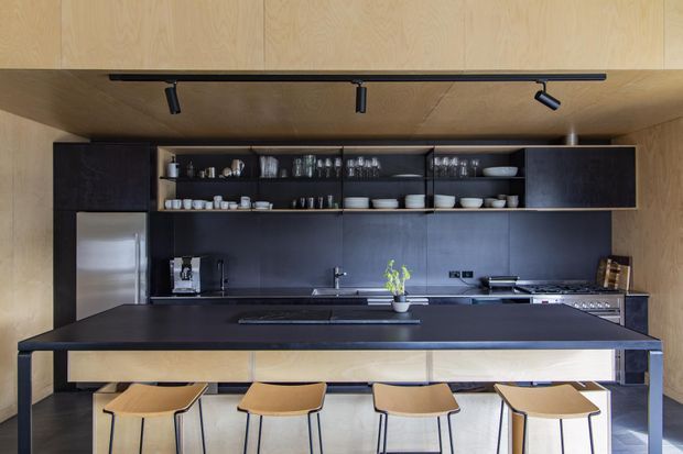 Contemporary open-plan kitchen with a large dark island and four timber bar stools. Full-height timber-look wall and ceiling panels surround dark built-in cabinetry, open shelving displaying crockery and glassware, integrated appliances, a sink, and black track lighting.