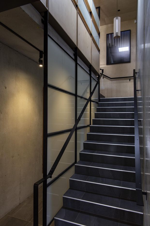 A narrow contemporary stairway rises through a tall interior void. The stairs are finished in dark charcoal tile with slim light grout lines and black metal handrails. One side features black-framed translucent glazing or panelled screens, while the surrounding walls combine raw concrete, pale timber wall panels and vertical textured cladding. A cylindrical pendant light hangs above, with a high-level window adding light to the stairwell.
