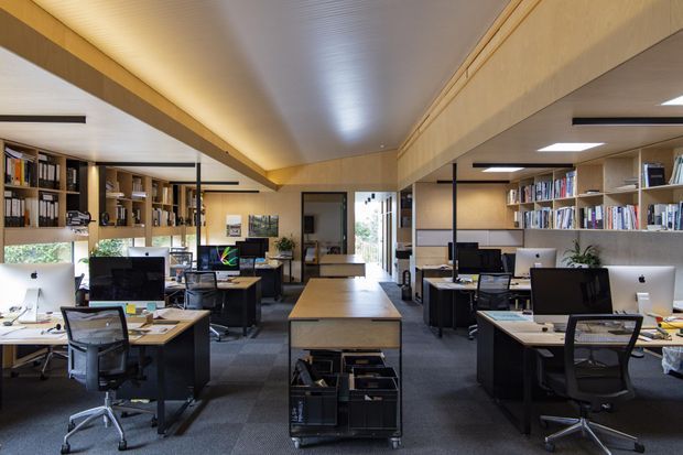 A bright, open-plan office interior with multiple workstations, desktop computers, ergonomic office chairs, extensive built-in bookshelves, and a central storage island. Warm timber-toned cabinetry and ceiling beams contrast with dark carpet flooring and black metal framing, creating a professional studio or workplace environment.